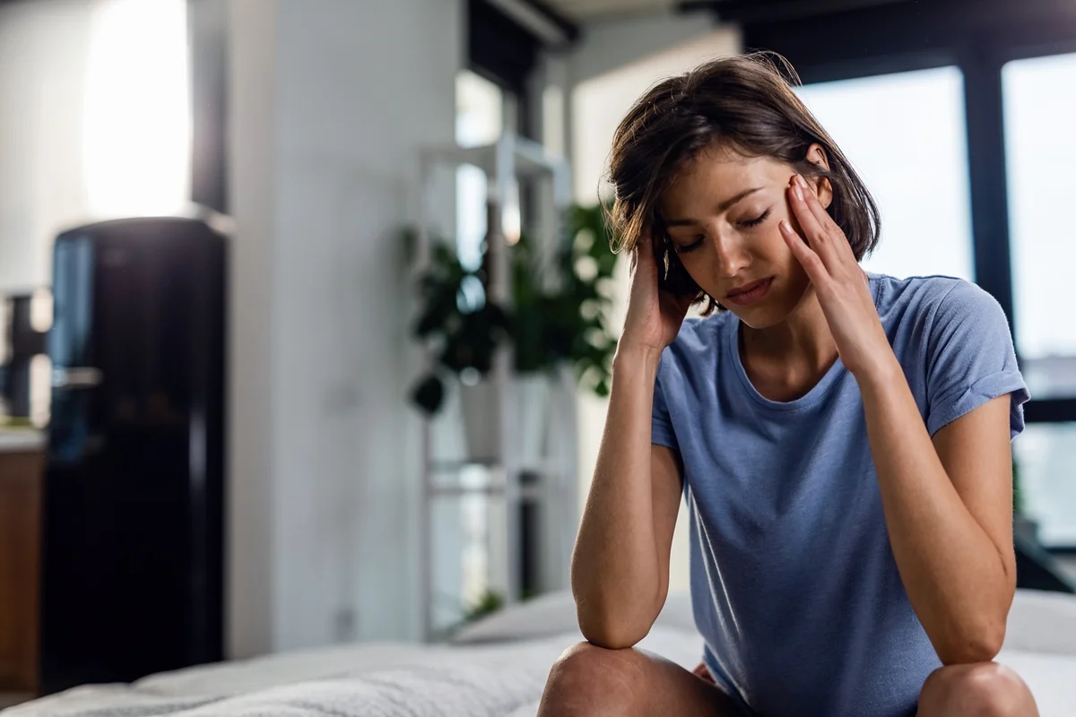 Mulher triste, sofrendo de dor de cabeça, sentada na cama com os olhos fechados enquanto segura a cabeça, sentindo dor. Esse pode ser um sinal de falta de vitamina b12 - Metrópoles